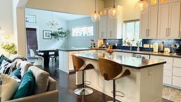 Una cocina moderna con armarios blancos, una isla central con taburetes, iluminación colgante y una zona de comedor con sofá y sillas.