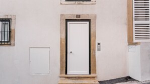 Une ruelle étroite, avec une porte blanche au fond, une petite fenêtre grillagée et une boîte électrique blanche fixée au mur.