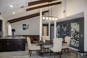 A dining area with a marble floor, a wooden dining table, and four upholstered chairs. There is a large floral painting on the wall and a modern pendant light above the table.