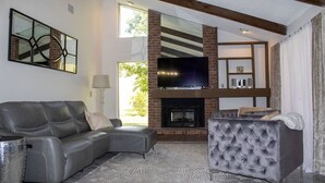 A modern living room with a leather sofa, a tufted armchair, a flat-screen TV above a brick fireplace, and a large window providing natural light. -  Private  Pool Home 
Nestled on the Golf Course (North Fort Myers)
