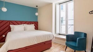 Une chambre à coucher avec un lit, une tête de lit rouge, une chaise bleue et une fenêtre donnant sur des immeubles.