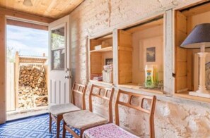 Interior rústico de madera con una puerta que da al exterior, un montón de leña y un banco con cojín estampado.