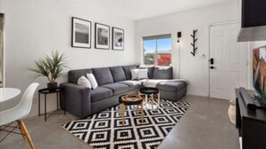 Een moderne woonkamer met een grijze modulaire bank, een ronde salontafel en een kamerkleed met een patroon. Er staat een eettafel met stoelen, een potplant en een televisie op een dressoir.