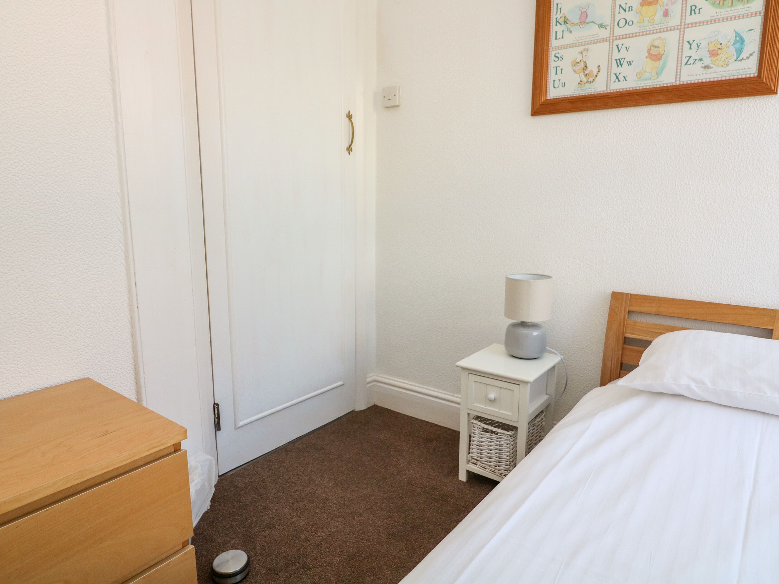 Quarto com penteadeira de madeira, mesa de cabeceira com luminária, cama com roupa de cama branca e um quadro com o alfabeto na parede.