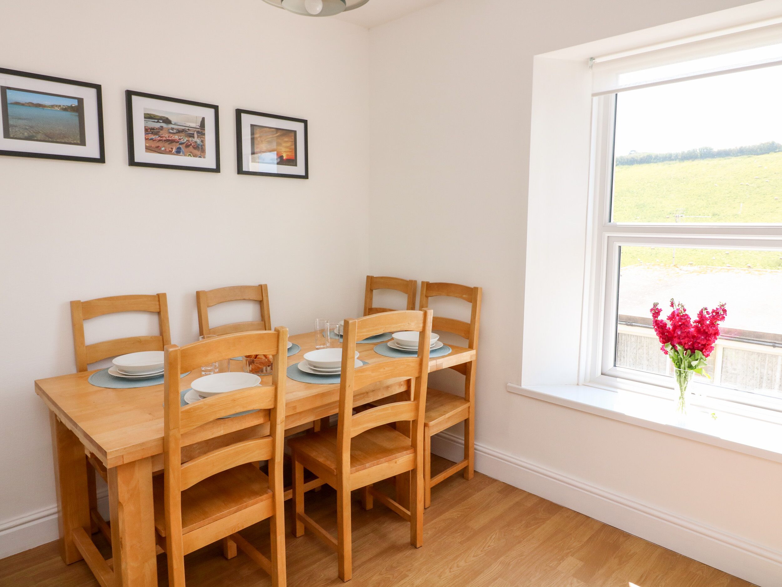 A dining area with a wooden table and chairs, framed pictures on the wall, and a window with a view of a field.