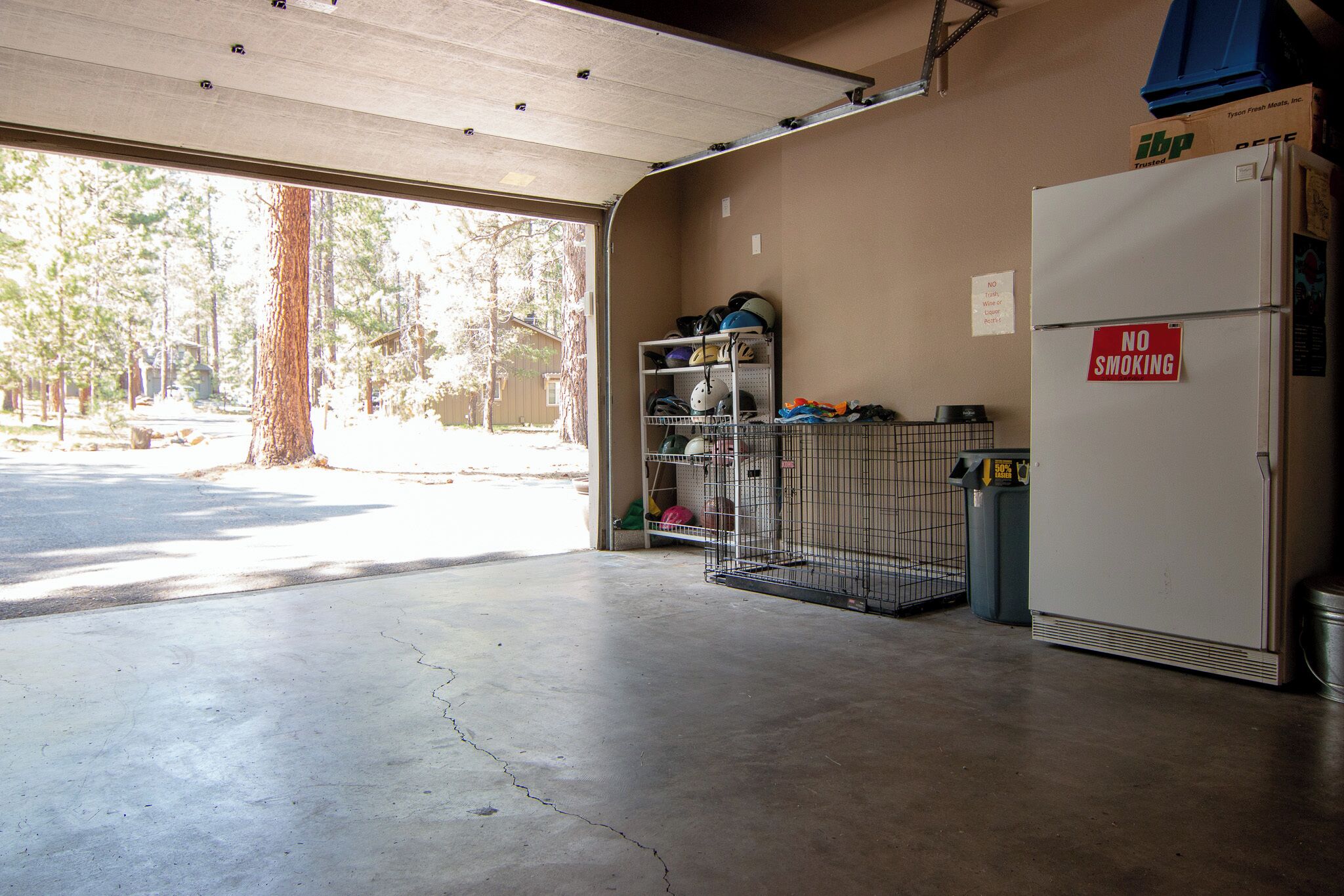 A garage with a concrete floor, a metal shelving unit with helmets and gear, a 'NO SMOKING' sign, and a refrigerator.