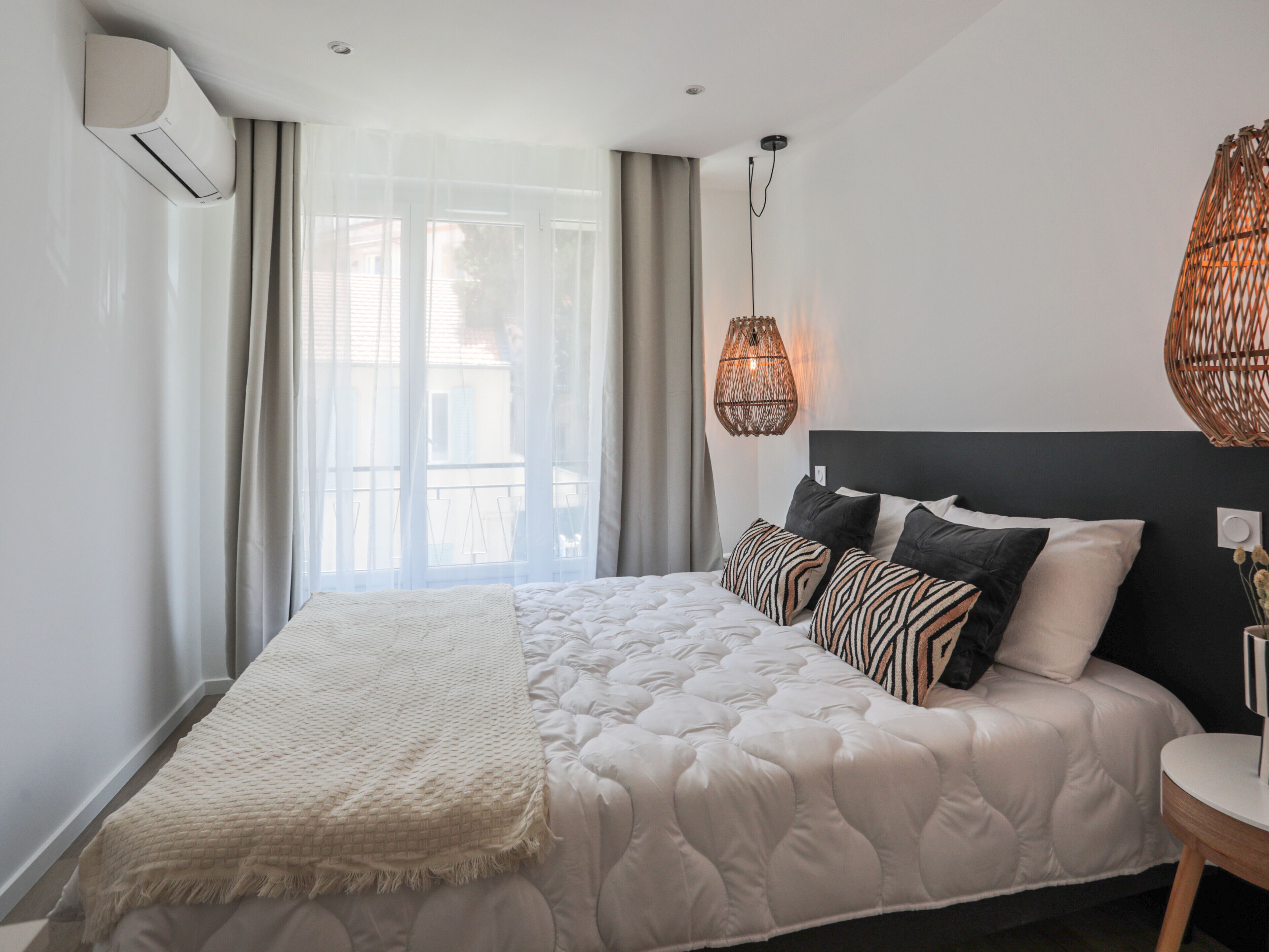 A modern bedroom with a large bed, a bedside table, a wall-mounted air conditioner, and a window with curtains.