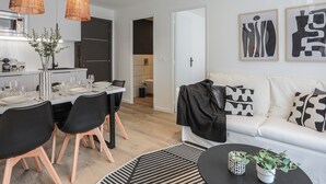 A modern living room with a white sofa, black and white patterned cushions, a black coffee table, and a dining area with a white table and black chairs. - Appartement Ebène - Welkey (Antibes)