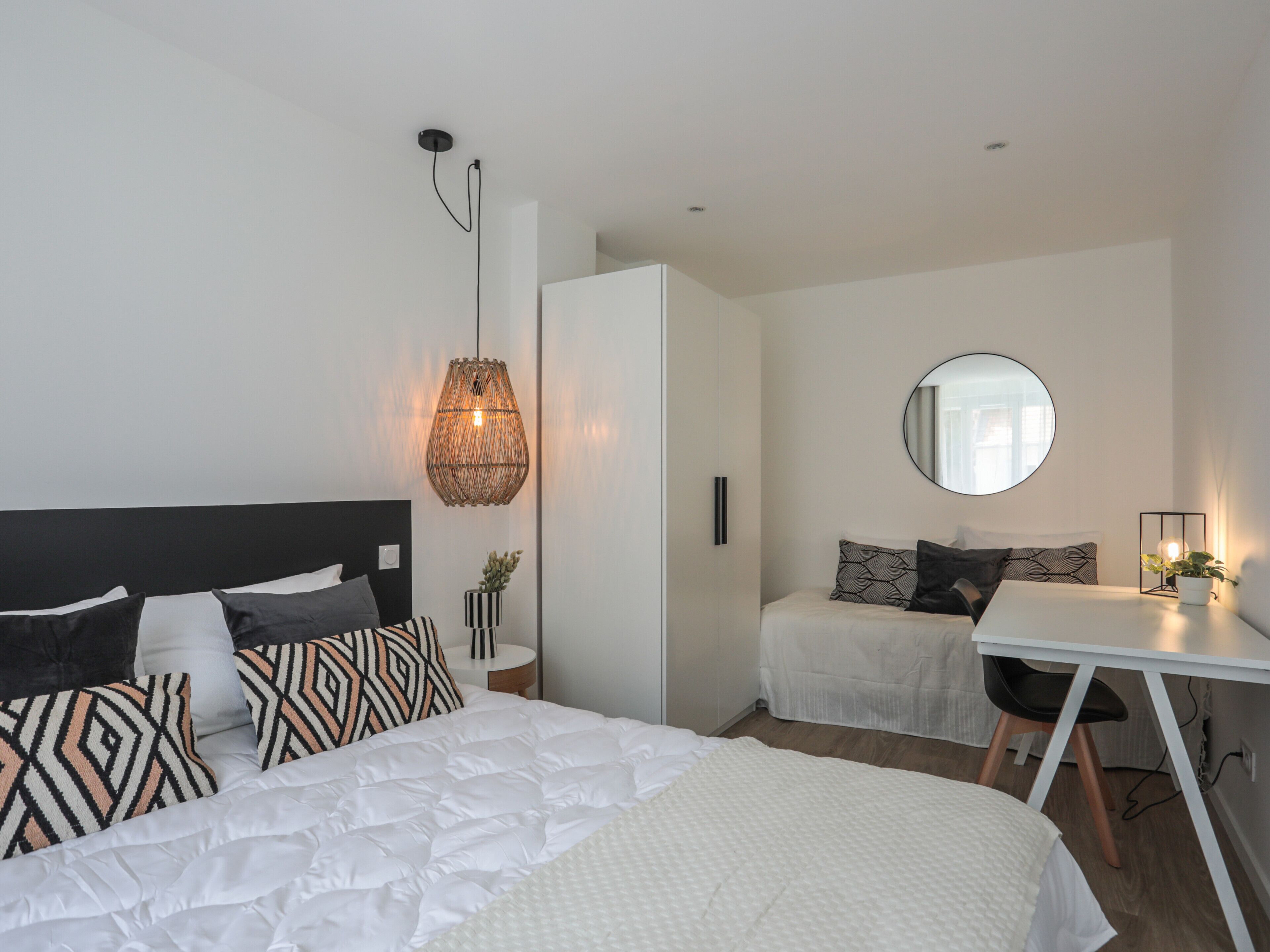 A modern bedroom with a large bed, a smaller bed, a desk, and a mirror.