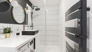 A modern bathroom with a white sink, black and white tiled walls, a built-in washing machine, and a shower with a black control panel. - Appartement Ebène - Welkey (Antibes)