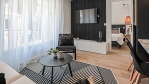 A modern living room with a sofa, a black armchair, a small round table with a plant, a black and white striped rug, and a TV mounted on a black wall. - Appartement Ebène - Welkey (Antibes)