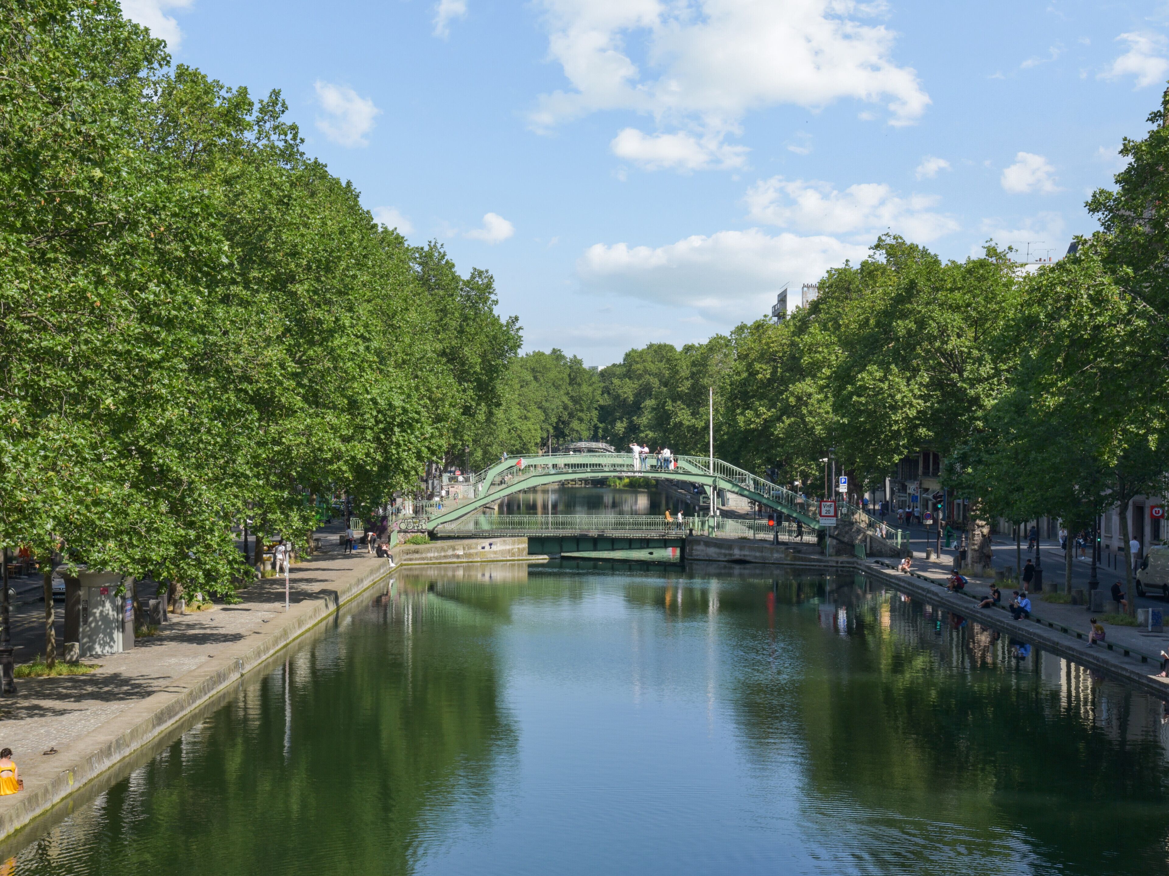 A green bridge over a calm river, surrounded by lush trees and a clear sky.