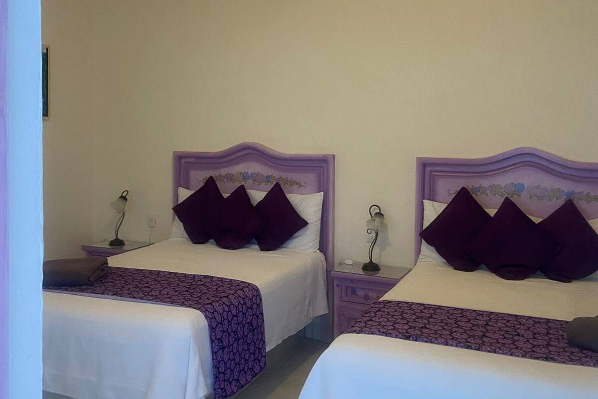 A hotel room with two beds, each with a purple headboard and white bedding. There are bedside tables with lamps and a small nightstand with a lamp.