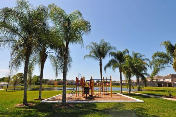 A playground with swings and climbing structures, surrounded by palm trees and a body of water, with houses in the background.