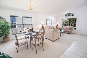 A spacious living room with a dining area, beige sofas, a chandelier, and large windows with views of greenery.