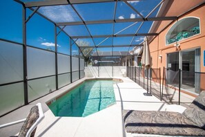 A covered outdoor swimming pool with a glass enclosure, a lounge area with a patterned sofa, and a glass door leading to a building.