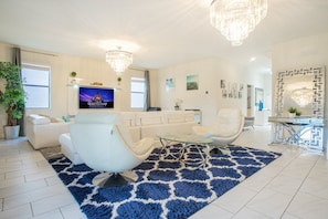 A modern living room with a flat-screen TV, white sofas, a glass coffee table, and a blue patterned rug.