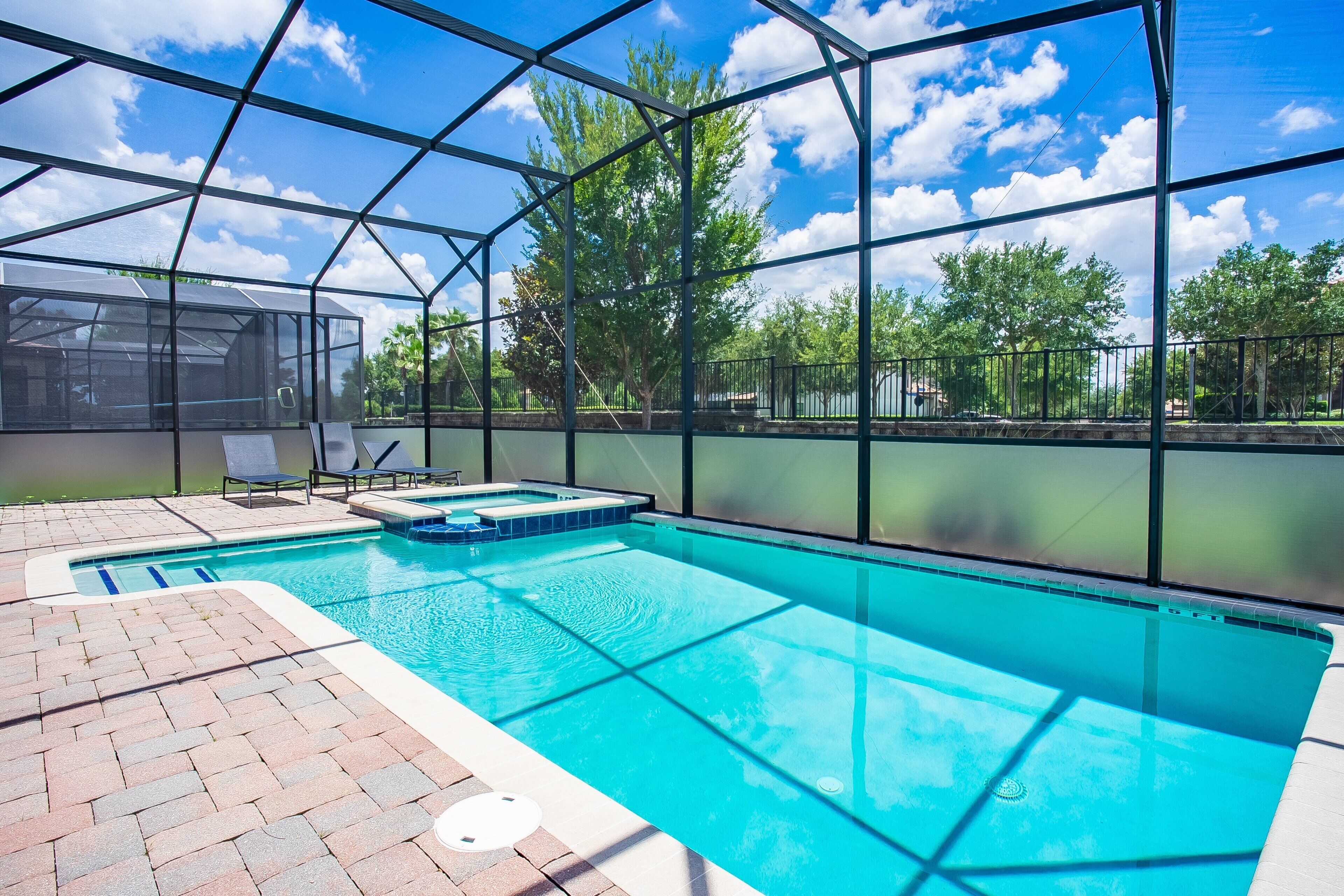 A swimming pool with a glass enclosure, surrounded by a brick patio and a metal fence.