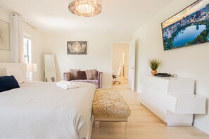 A bedroom with a bed, a dresser, a bench, a chandelier, and a painting of a cityscape.