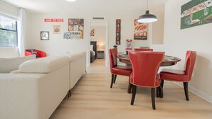 Ein moderner Wohnbereich mit Essplatz, der einen runden Tisch und rote Stühle, ein weißes Sofa sowie unterschiedliche Wanddekorationen, darunter Filmplakate, umfasst.
