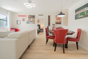 Ein moderner Wohnbereich mit Essplatz, der einen runden Tisch und rote Stühle, ein weißes Sofa sowie unterschiedliche Wanddekorationen, darunter Filmplakate, umfasst.