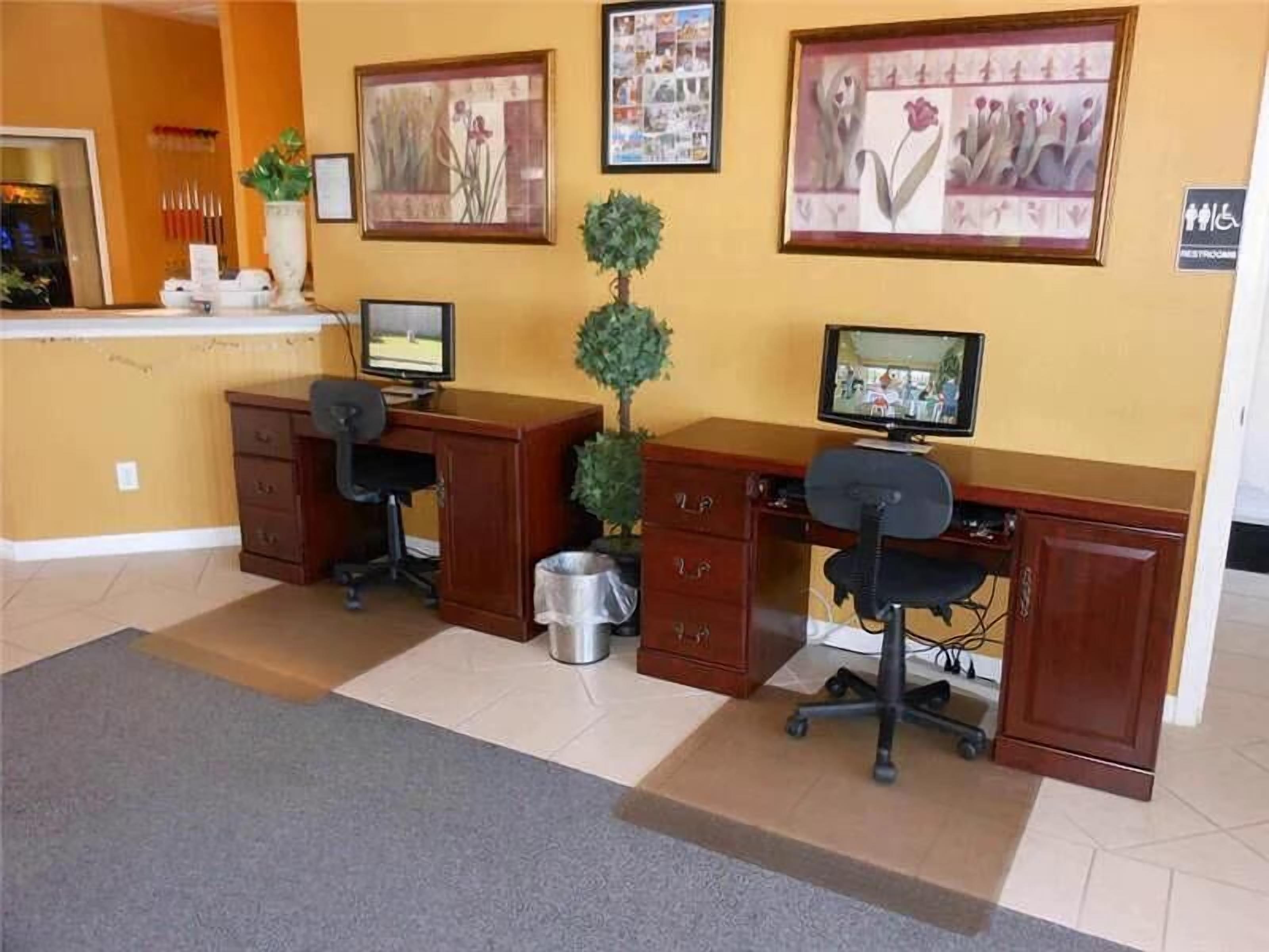 Zona de recepción con dos mostradores de madera, cada uno con un monitor de computadora, una silla y una pequeña planta en maceta. Hay cuadros en la pared, un bote de basura y un letrero que indica el baño.