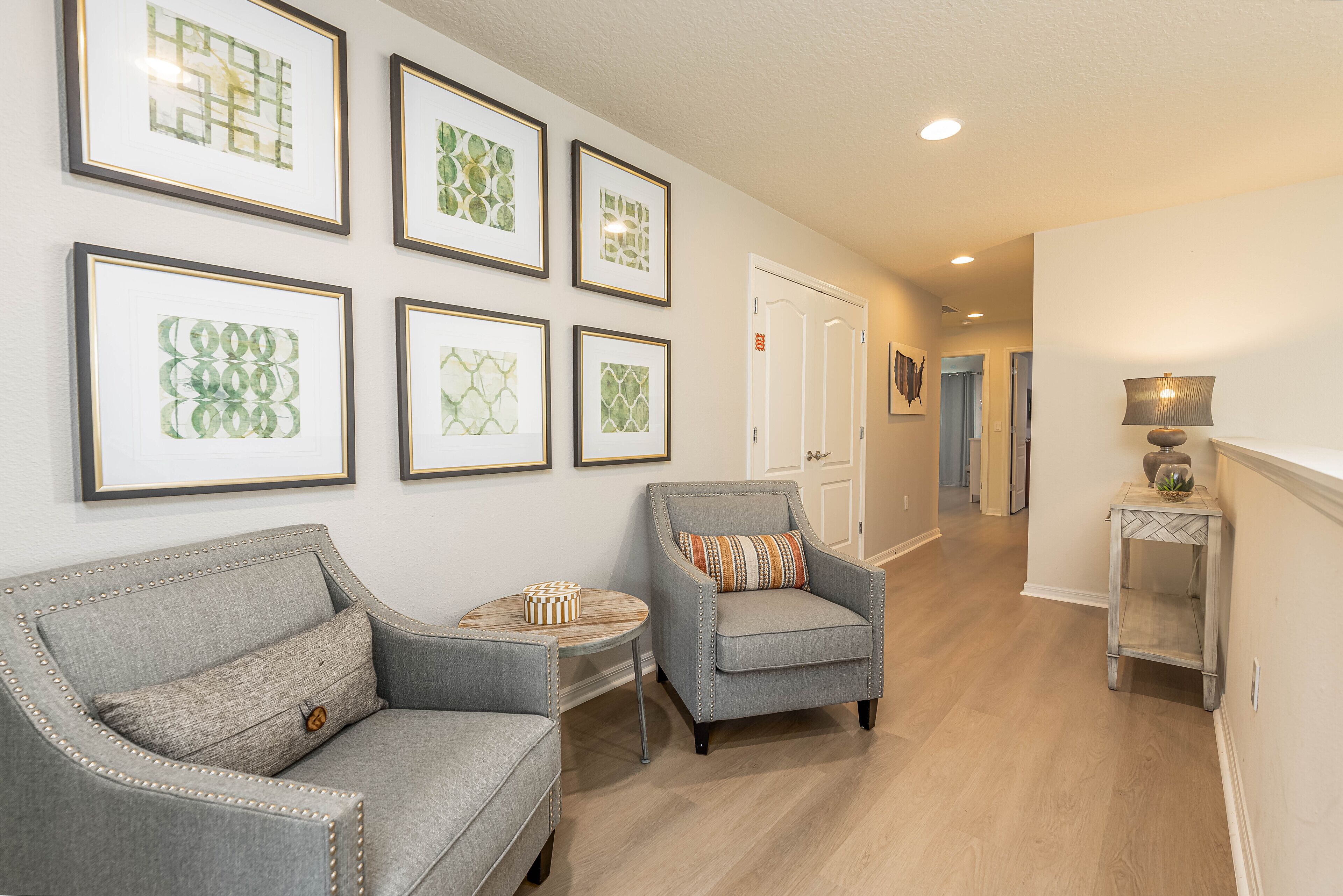 A hallway with a grey armchair, a small round table, and a side table with a lamp. There are framed pictures on the wall and a door at the end of the hallway.