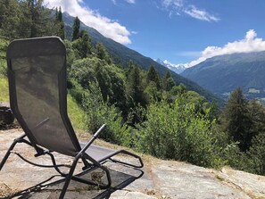 A chair on a stone ledge overlooking a mountainous landscape with dense greenery and a clear sky. - Outside: Apartment in a chalet for 2 people with balcony, terrace with garden furniture,  a breathta (St-Luc)