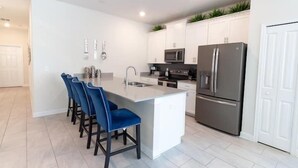 A modern kitchen with a central island, bar stools, white cabinets, stainless steel appliances, and a tiled floor. - Spacious 5-bedroom townhouse in Kissimmee, perfect for family getaways! (Kissimmee)