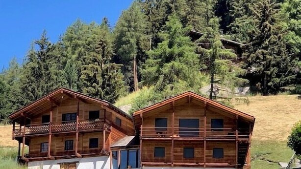 Dos cabañas de madera con balcones, ubicadas en un área cubierta de césped con pinos al fondo.