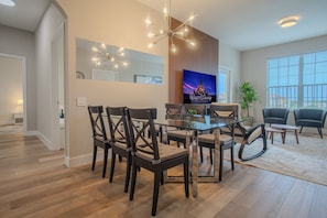 A modern living room with a glass dining table, dark wooden chairs, a large flat-screen TV, and a potted plant.