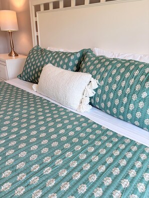 Un lit avec une couvre-lit à motifs floraux verts et des oreillers blancs, une lampe de chevet et une tête de lit blanche.