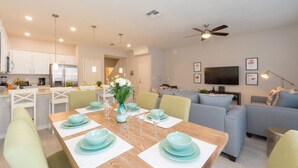 A modern kitchen with white cabinets, a dining area with a wooden table set for four, and a living room with a grey sofa and a flat-screen TV.