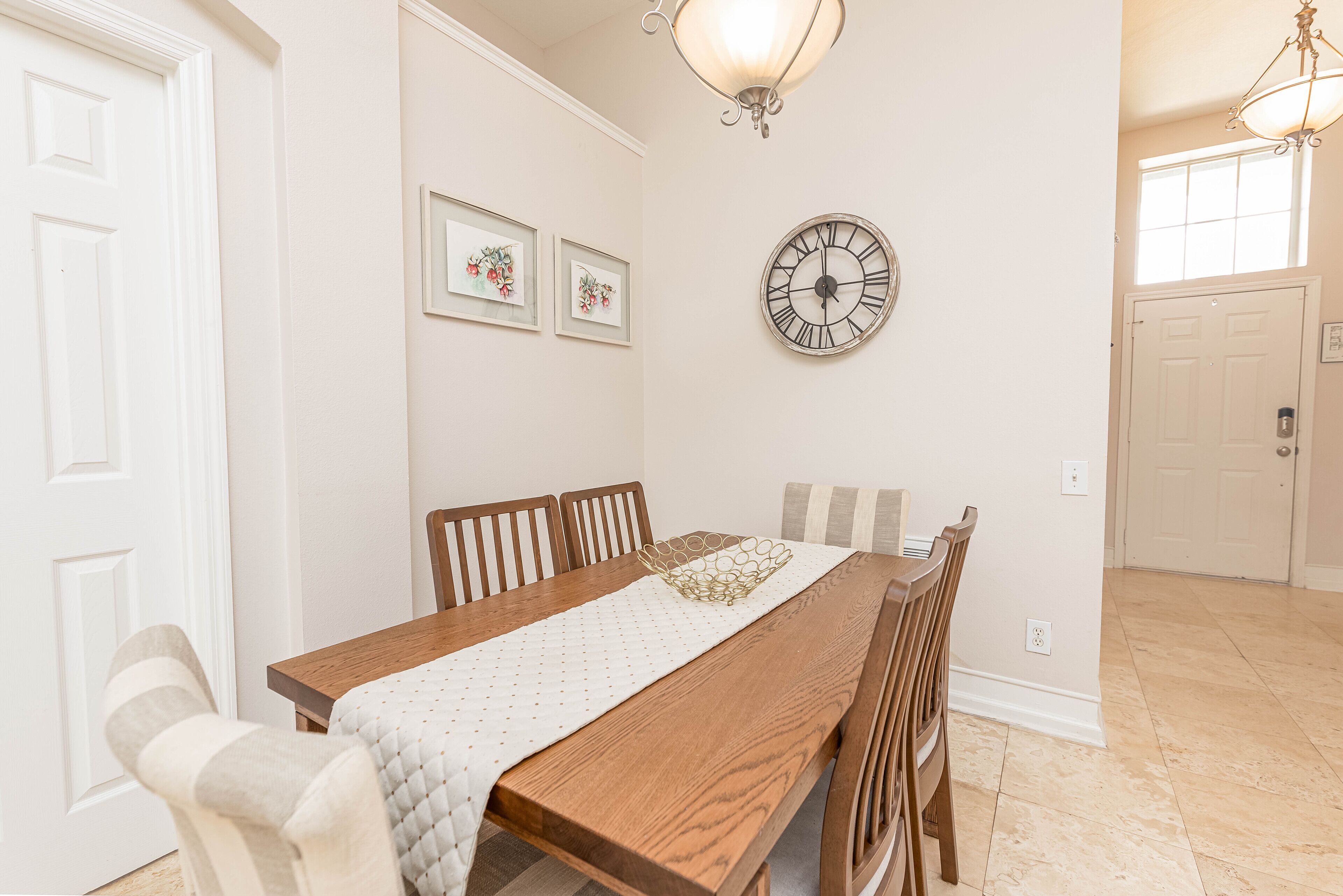 A dining room with a wooden table, chairs, a wall clock, and framed artwork.