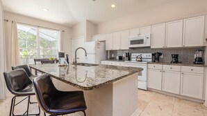 A modern kitchen with white cabinets, a granite countertop island, black bar stools, and a large window. - Spacious 4-bedroom home in Kissimmee, perfect for family getaways! (Kissimmee)