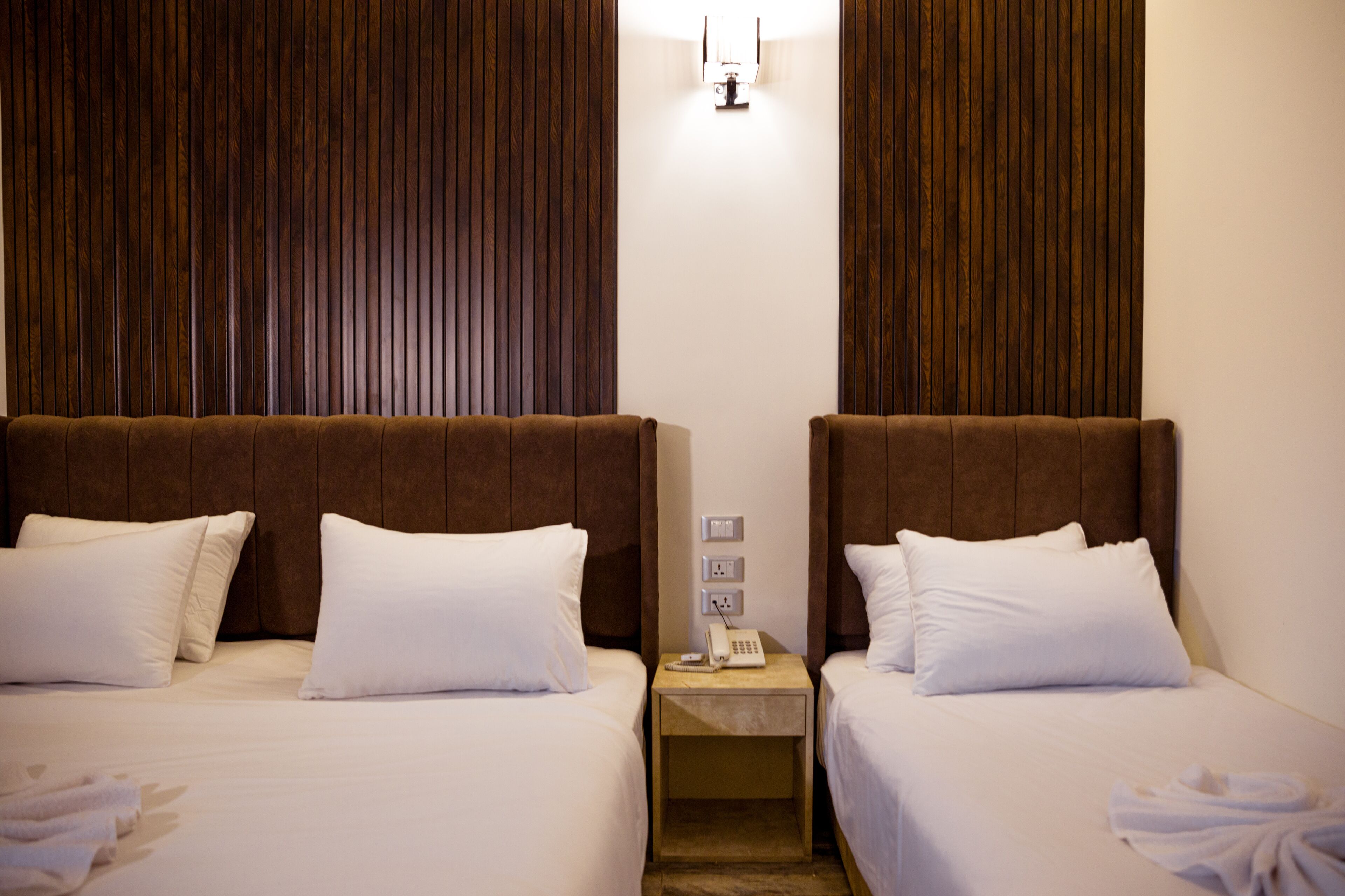 A hotel room with two beds, each with white bedding and brown headboards. There is a nightstand between the beds with a telephone and a light switch.