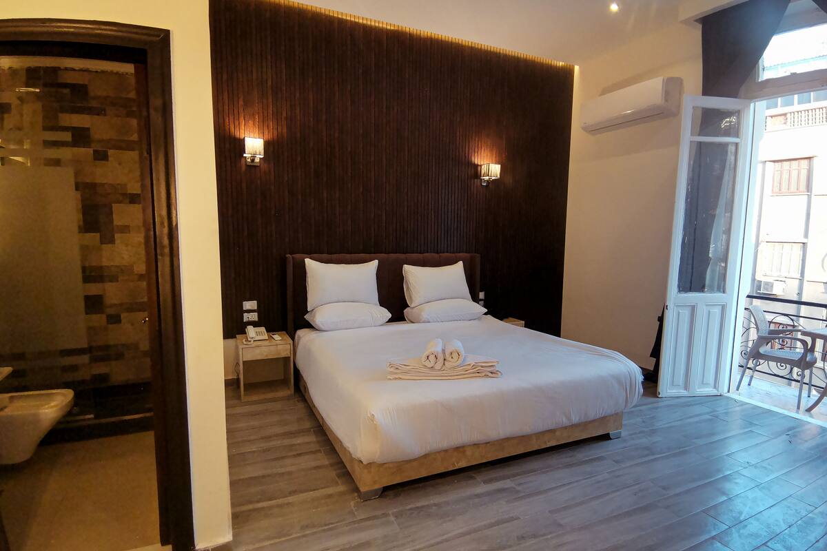A hotel room with a large bed, two bedside lamps, a wooden headboard, and a balcony with a chair.