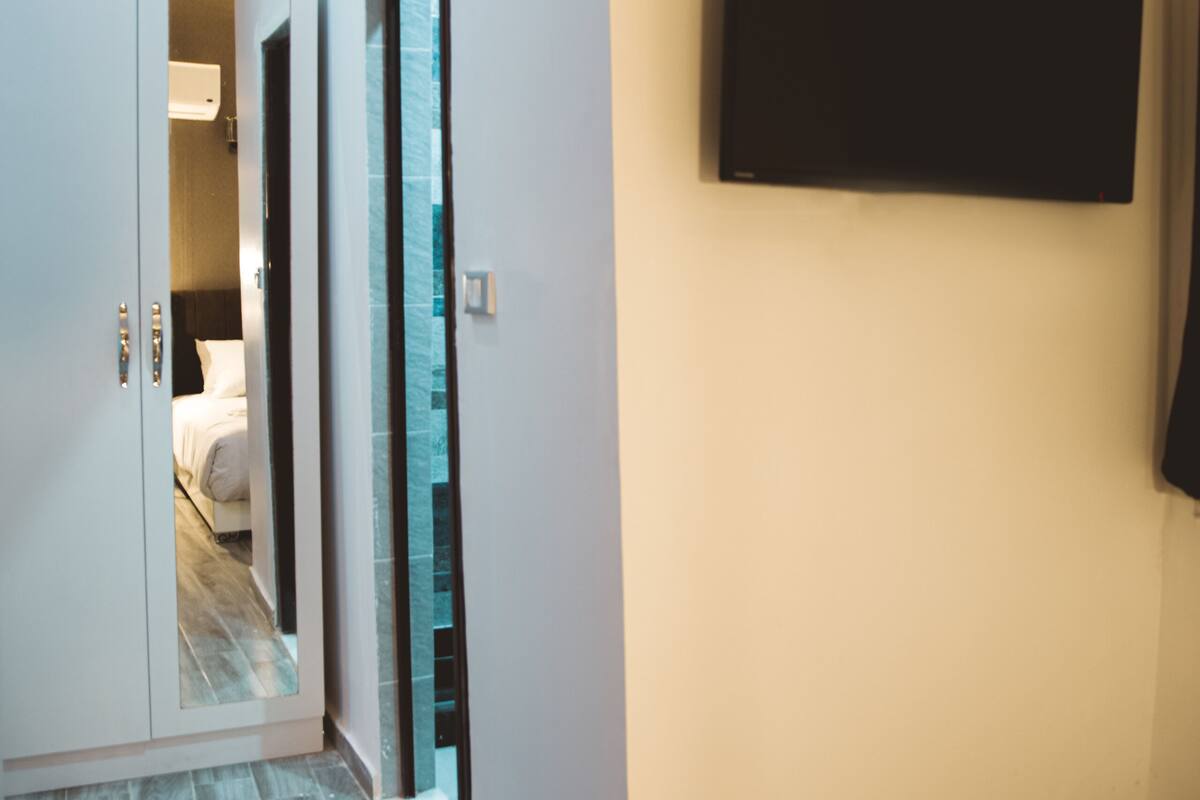 A hotel hallway with a wooden floor, a bed in the background, and a flat-screen TV mounted on the wall.