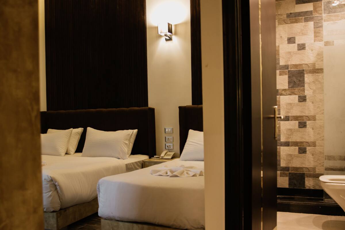 A hotel room with two beds, a wooden headboard, a wall-mounted light, and a bathroom with a tiled shower wall.