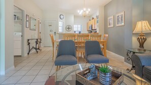 A spacious living area with a glass coffee table, two blue armchairs, a dining table with chairs, a kitchen in the background, and a clock on the wall. - Charming 3-bedroom townhome in Kissimmee, perfect for family getaways! (Kissimmee)