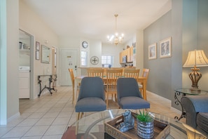 A spacious living area with a glass coffee table, two blue armchairs, a dining table with chairs, a kitchen in the background, and a clock on the wall. - Charming 3-bedroom townhome in Kissimmee, perfect for family getaways! (Kissimmee)