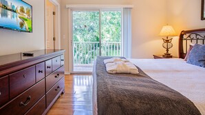 Een slaapkamer met een groot bed, een dressoir, een televisie, een balkon met uitzicht op groen en een lamp.