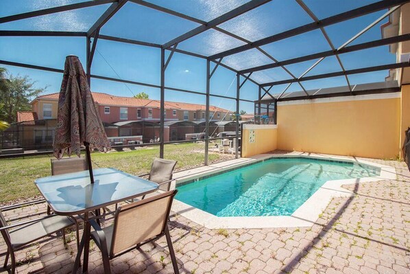 Una piscina exterior cubierta con mesa y sillas de vidrio, rodeada por un patio de ladrillo.