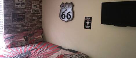 A bedroom with a bed, a brick wall, a Route 66 sign, a Ford parking sign, and a television.