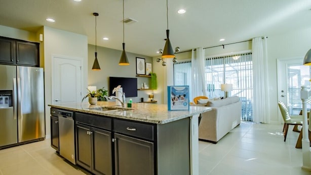 Una cocina moderna con una isla central, electrodomésticos de acero inoxidable, iluminación colgante y vistas a una sala de estar con un sofá y una mesa de comedor.