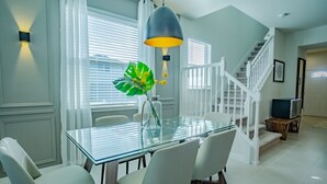 Un comedor con una mesa de vidrio, sillas blancas, una lámpara colgante y una escalera al fondo.