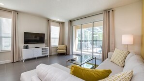 Un salon moderne avec un canapé blanc, une table basse en verre, un téléviseur à écran plat posé sur un meuble blanc, et une vue sur l’extérieur grâce à de grandes fenêtres.