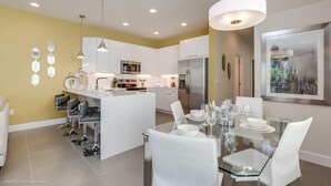 Une cuisine moderne avec des meubles blancs, des appareils en acier inoxydable et un îlot central. On y trouve également un coin repas avec une table en verre et des chaises blanches.