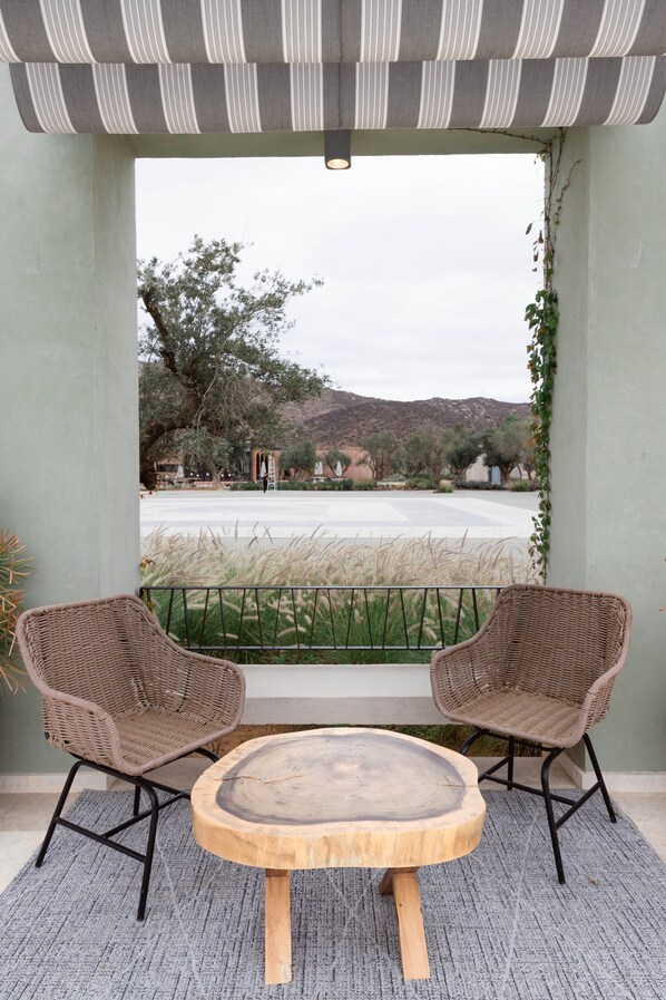 A balcony with wicker chairs and a round wooden table, overlooking a landscaped area with trees and a distant view of hills. - Hotel Andana Resort (Valle de Guadalupe)