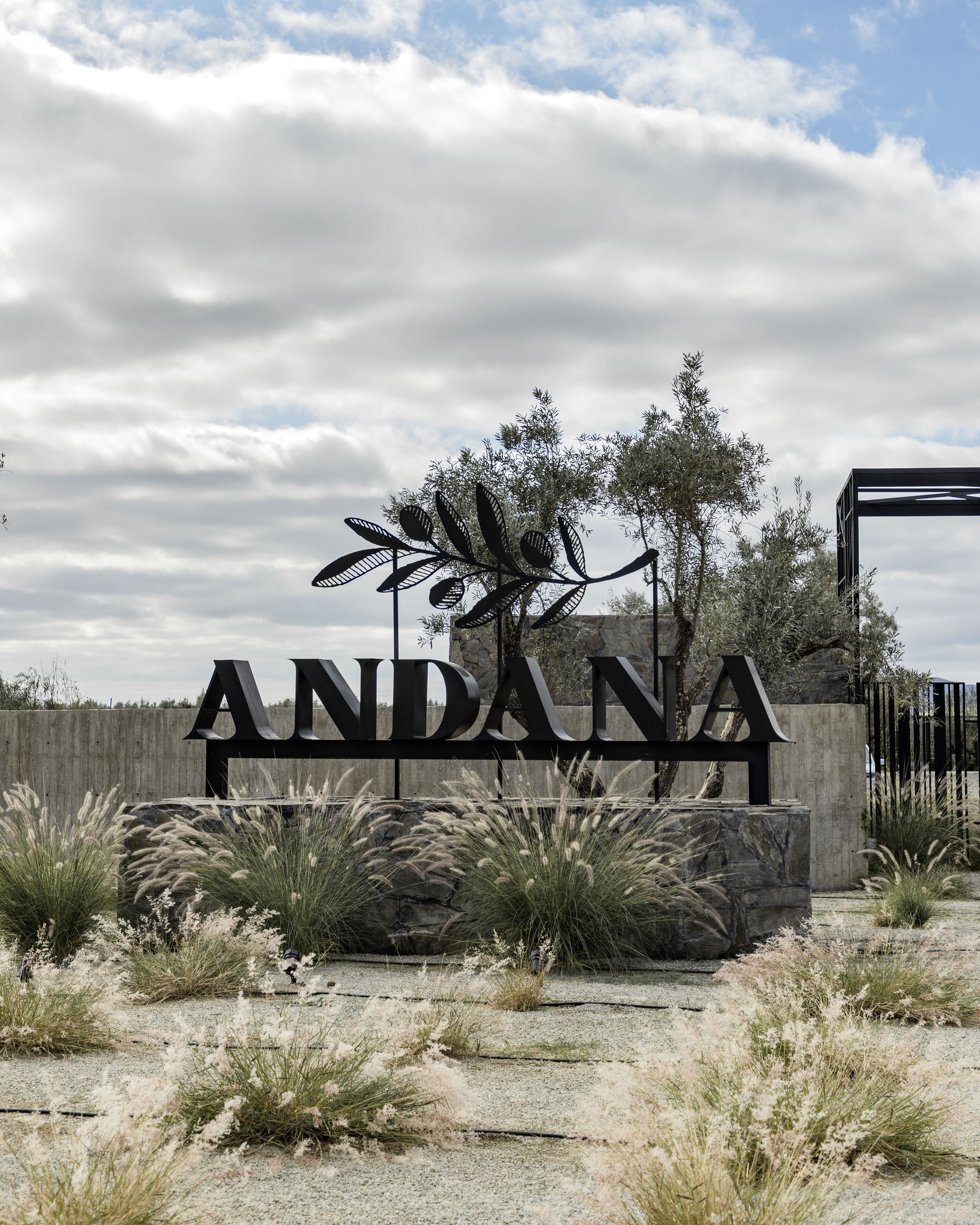 Un letrero con la palabra 'ANDANA' en letras grandes, rodeado de follaje decorativo y un muro de piedra.